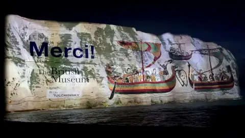 White cliffs of Dover at night, covered in a giant projection with images of wooden boats from the tapestry, and "Merci"