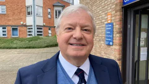 Councillor Alan Amos, wearing a blue suit and blue jumper, standing outside the civic centre in Pershore.