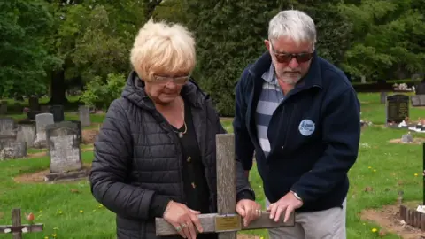 Liz and Malcolm Hare are standing at Pete Wilson's grave with their hands on the wooden cross. They are both looking down at the grave. 