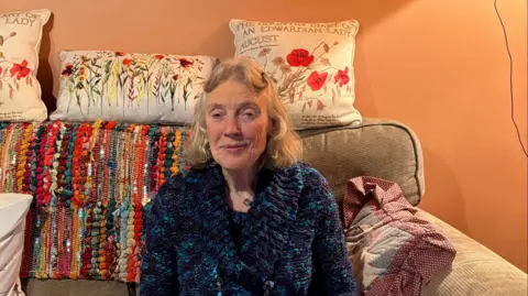 Annette Turto is sitting on a grey sofa. She is wearing a blue coat. The sofa has lots of pillows.