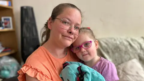 Kelly has tied back dark hair. She is wearing glasses and has nose piercings. She is wearing a coral coloured t-shirt with ruffled sleeves. Her daughter Connie is sitting on her lap and their faces are close together. Connie has her hair tied back and is wearing pink framed glasses. She is holding a blue soft toy.