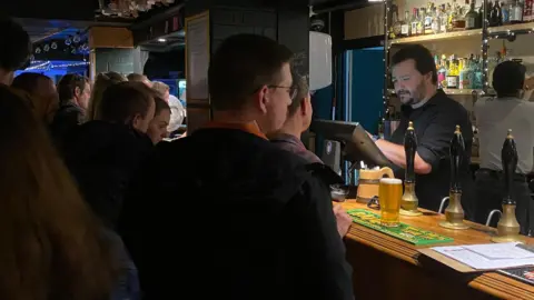 A crowd of people ordering drinks at a bar. A man in a black shirt works the till behind the bar.