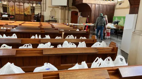 Dozens of food parcels in carrier bags line wooden church pews.