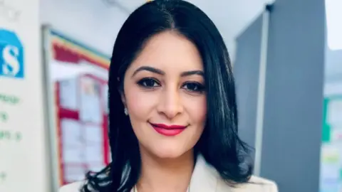 Shabina Asad Qayyum Shabina wearing a white blazer - smiling for the camera. She has black shoulder length hair and is standing indoors.