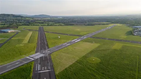 A small airfield shown from above, with two runways crossing. Countryside can be seen in the distance, and there are no planes on the tarmac. 