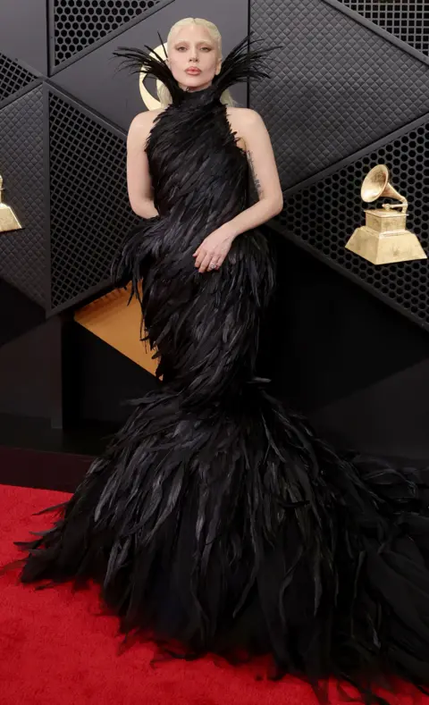 Getty Images Lady Gaga luce un vestido negro largo con un diseño de plumas mientras posa para una foto en la alfombra roja de los Premios Grammy.