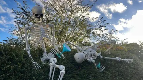 Two plastic skeletons are on a large hedge. One is sitting upright and holding a blue plastic cocktail glass. The other is lying down with a plastic glass next to it.