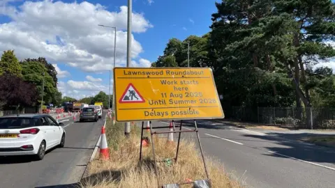 Sofia Luis-Hobbs/BBC : A yellow roadworks sign beside a coned-off carriageway warns of construction at Lawnswood Roundabout. The sign reads: “Lawnswood Roundabout. Work starts here. 11 Aug 2025 until Summer 2026. Delays possible.” Cars travel along the road past traffic management measures, with trees, streetlights and ongoing roadworks visible under a bright sky with scattered clouds.