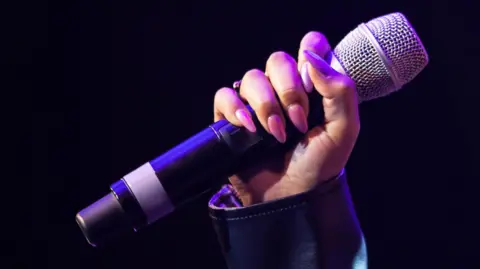 A hand, with pink painted nails, holding a microphone. The background of the image is black. 