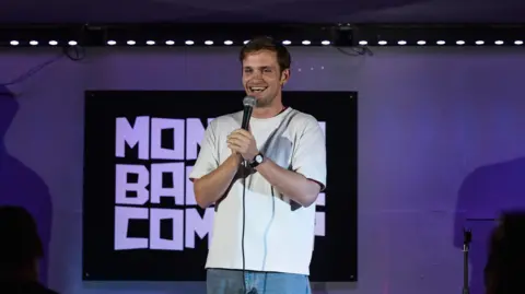 Andrew Jackson Vittorio Angelone stands on stage in a white t-shirt, holding a microphone