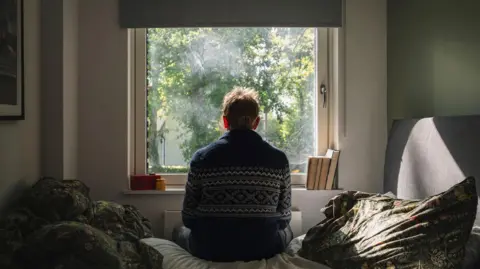 A man is seen from behind as he is sat on a bed looking out onto a sunny day.