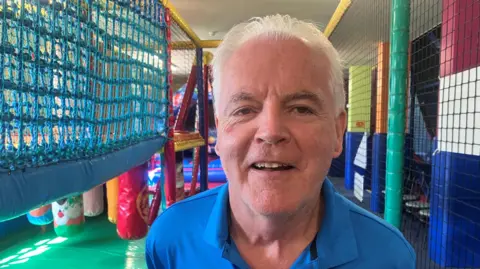 Michael Hazzard is wearing a blue polo shirt and has grey hair. He stands in a soft play area at Coco's Adventure Playground, surrounded by climbing frames, netting and children's play equipment.