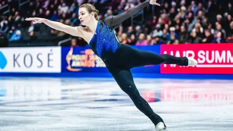 Getty Images Kristen Spours competing for Great Britain on the ice. She is mid-routine, with one leg raised behind her in the air. Both of her arms are raised above her head and she is leaning. She is wearing a black and bright blue costume and white skates. The crowd is blurred behind her.
