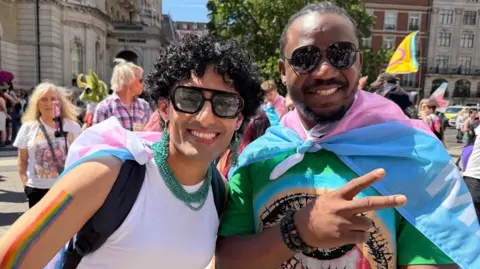 A photo of Ansar, on the left, who appears to be in their 20s or 30s and has short black curly hair and wears black aviator sunglasses, a white vest top and a green beaded necklace and earrings. They have a transgender pride flag tied to their backpack and are standing next to David, who appears to be about the same age and who is smiling, also wearing aviators and a trans pride flag as a cape.