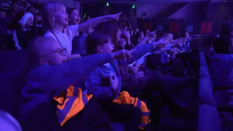 Children from Fieldhead Primary Academy in Kirklees are screaming and shouting and all pointing at the stage in a theatre.