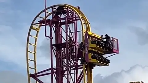 Supplied A ride operator stands on a moveable platform next to the roller coaster cart, which is near a high point on the track. The yellow cart, which is facing upwards, has two rows of four black seats. The operator is helping someone out of the cart