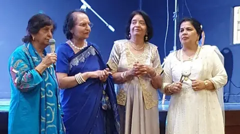 Essex Asian Women's Association Four women speaking to a crowd. One is holding a microphone while the other three smile on.