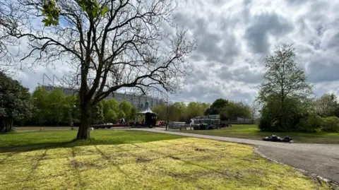 A view of Leazes Park in the spring, with St James' Park in the background. There is an expanse of lawn, several trees and a bandstand.