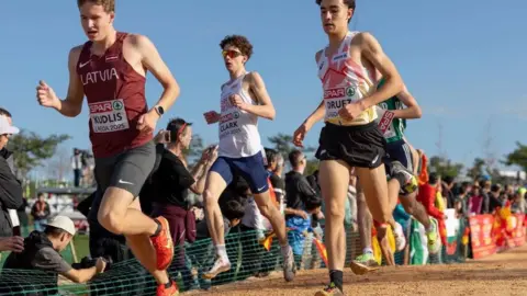 sammellishphoto Three runners, including Michael Clark, are wearing shorts and vests while running on a sandy stretch of the race track. Supporters are watching them from behind a green netted barrier.