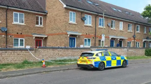 A police car parked on Saunders Road in Oxford, with police tape surrounding a patch of grass in front of a wall, with a row of houses in the background 