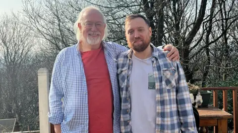 Supplied Two men stand side by side outside on decking, with trees in the background. One is wearing glasses, and has white hair and a beard. The other has brown hair and a beard. Both are smiling, and both are wearing a T-shirt underneath a chequered shirt.