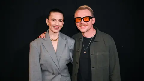 Hayley Atwell in a grey suit and Simon Pegg in a green shirt smile with their arms around each other