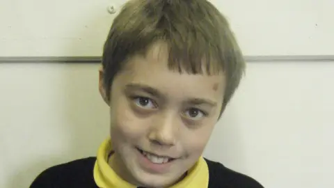 Toby Hayward Toby, as a child, in a school uniform, with a yellow top and black jumper. He has short hair and is smiling and looking straight at the camera. 
