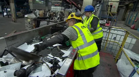 Two workers in hi-vis vests, safety gloves, goggles and hard hats sort through electrical waste on a conveyer belt in a factory.