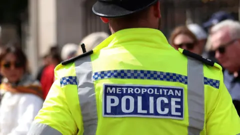 A Met Police officer with his back to the camera stands on a London street. There are passers-by slightly out of focus.