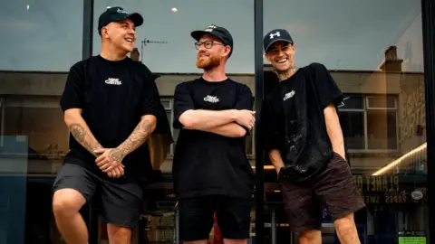 Simon Hogarth Three Bone Cartel staff members outside the shop in black t-shirts with white letter branding and in black shorts and black caps. Simon Hogarth is in the middle and has a red beard