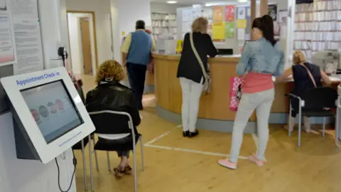 People stand and sit in a doctor's surgery waiting room. On the left is a white screen to check in for appointments. In the right is a reception desk