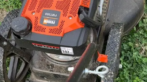 Fenland Orchards Project A close-up of an orange and black mower, showing the wheels and the motor. A logo on the mower includes the name "Feider Machines".