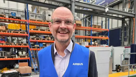 Simon Galpin in an Airbus factory. There are high orange shelves behind him in the large warehouse. He is smiling at the camera and is wearing a blue vest that says Airbus on it, over a suit.