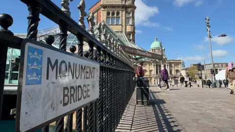 Kevin Shoesmith/BBC A bridge over a former dock. A sign reading Monument Bridge is fixed to railings next to the water. In the distance is City Hall with its lead, domed roof.