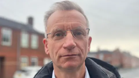 A man looking at the camera, he has light hair and glasses.