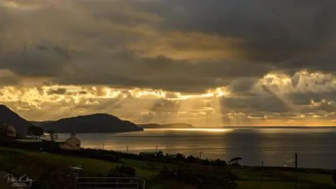 Sunset at Niarbyl Bay on a cloudy day. The remaining yellow light is streaming through the clouds illuminating part of the water below. A silhouette of the Calf of Man in the distance.