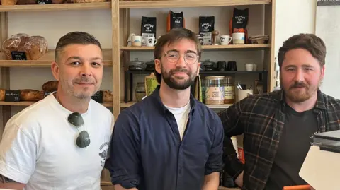 Grace Wood/BBC Three men stand behind the counter in a cafe smiling at the camera