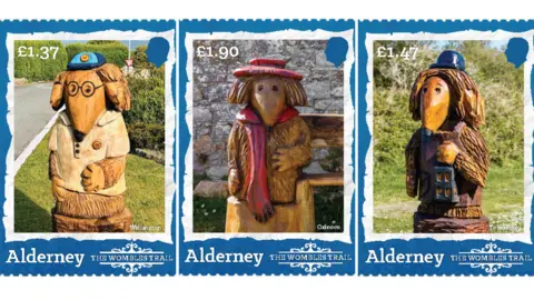 Guernsey Post Three of the stamps. On the left is the £1.37 stamp which shows a Womble sculpture wearing a blue cap, white T-shirt and pair of glasses. It has shoulder-length hair and a pointed nose and is standing in a planted and lawn area. The middle stamp is a £1.90 one which shows a Womble sculpture wearing a red scarf and hat sitting on a bench with a stone wall behind it. The one on the right is the £1.47 stamp. It shows a Womble wearing a blue hat and holding a lantern in a grassy area with hedges behind it. It has shoulder-length hair.
Each stamp features the silhouette of King Charles III's head and the wording: Alderney: The Wombles Trail.