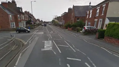 A view of a road. A black car is pulling out at a junction up ahead on the left and there are rows of houses either side of the road.