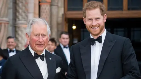 Head and shoulders pics of then Prince Charles and Prince Harry in London in 2019. They are both wearing tuxedos and smiling at the camera. Harry is on the right
