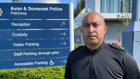 BBC Image Superintendent Deepak Kenth is standing in front of an Avon and Somerset Police sign. Deepak has very short hair and is wearing police uniform. 