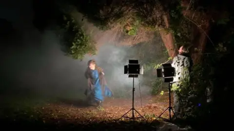 Mike Alsford and Darren Miles An actress in Victorian clothing running through a dark park. She is illuminated by stage lights and a member of the film crew is standing behind the lights.