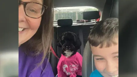 Julie Blackburn A selfie of a woman with long brown hair and glasses, a black and white dog in a pink Cancer Research UK T shirt and a boy in a blue T shirt. They are sitting in a car. 