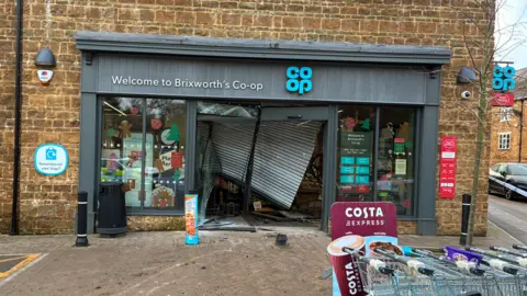 A shop with a grey exterior and a blue "Co-op" sign. The front of the store is severely damaged, with the main door smashed and debris on the ground.