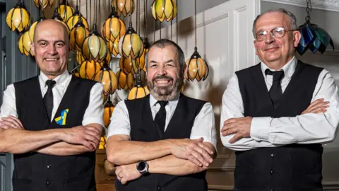 Jean Paul, Nelson and David all wearing white shirts, black ties and black waistcoats with their arms folded standing in a bar.