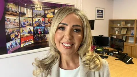 Noella Murphy. She has long blonde hair, wearing a cream blazer and white top.