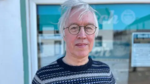 Brian Palmer stands in front of an office for the newspaper The Ileach - a sign for which can be seen in the window behind him. Brian wears a Nordic knitted jumper in dark blues and grey - he has white hair, which is blowing in the wind, and round glasses.
