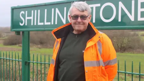 A smiling Gavin Collins standing in front of one of Shillingstone's green station signs. Behind him is countryside. He is wearing glasses and an orange hi-viz coat.