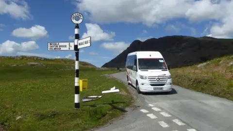 Lake District National Park Authority A white minibus prepares to turn right at a junction on a narrow Lake District road. Mountains can be seen in the background. A road sign gives the distances to Wasdale and other nearby locations.