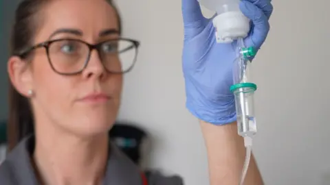 MARTIN GILES/BBC Matron Lana Goodwin holds up an intravenous drug drip. She has blue surgical rubber gloves on. Ms Goodwin is wearing brown glasses, has her hair tied back and is wearing a grey uniform with red seams.